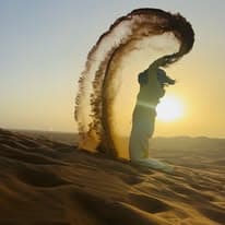 Guest silhouetted against the low desert sun, throwing an arc of golden sand from a dune crest